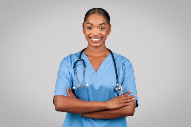 Gülümseyen, kendine güvenen, bin yıllık doktor cerrahı, göğsünde çapraz kollar olan mavi üniformalı hemşire, gri stüdyo arka planında izole edilmiş. Tıbbi reklam ve teklif, sağlık hizmetleri, profesyonel yardım, danışmanlık