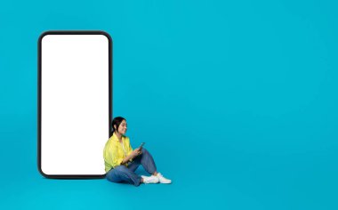 Sosyal ağlarda akıllı telefonlarda sohbet eden mutlu Çinli kadın, mavi stüdyo arka planında izole edilmiş, boş ekranlı büyük telefonun yanında oturuyor. Çevrimiçi iletişim için uygulama, reklam ve teklif, satış