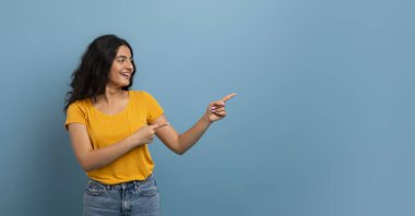 Sarı tişört ve kot pantolon giyen mutlu, kıvırcık, genç Hintli kadın reklam ve gülümseme için fotokopi alanını işaret ediyor. Mavi stüdyo arka planında izole edilmiş, panorama.