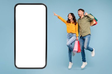 Genç Avrupalı eşler alışveriş çantalarıyla zıplıyorlar, kadınlar boş ekranlı dev cep telefonlarını işaret ediyor, mavi stüdyo arka planında satış reklamları için mükemmel.