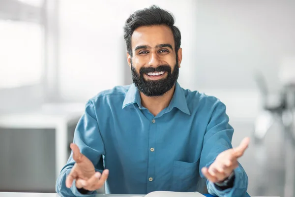 Happy bearded indian man with wide smile wearing blue shirt extends his hands with welcoming gesture while sitting at desk in office environment, having video call