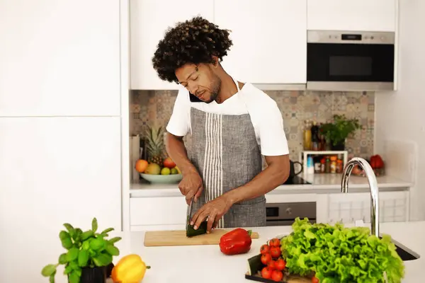 Young African American man chats on his smartphone while chopping vegetables in modern kitchen indoor, communicating and preparing healthy lunch. Nutrition concept