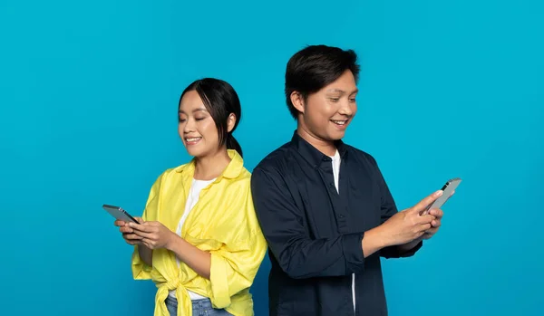 Cheerful chinese man in casual and woman chatting at smartphone at social networks, isolated on blue studio background. App for online shopping, communication, ad and offer