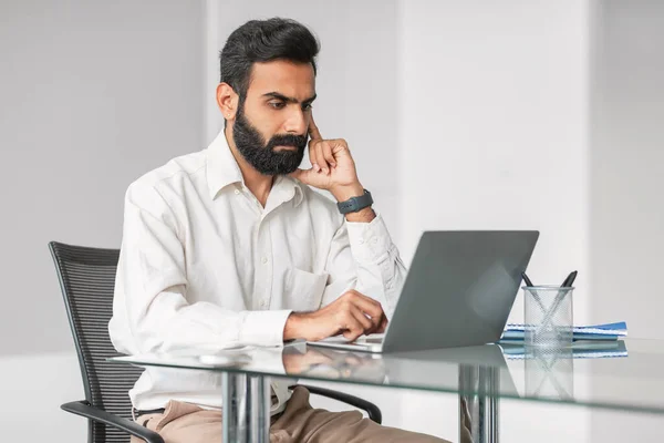 Deeply concentrated indian professional with beard diligently working on laptop at glass office table, showing dedication and attentiveness to his task