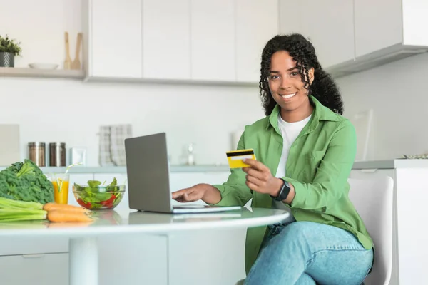 African American woman shopping ordering groceries online with credit card and laptop at home kitchen, sitting at table and smiling to camera. Financial savvy and modern lifestyle