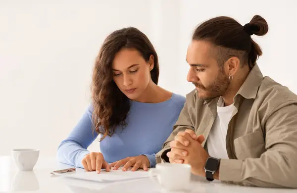 Loving couple looking at papers, carefully studying documents sitting at desk indoors, reading terms and conditions, reviewing agreement, considering mortgage loan offer, personal insurance