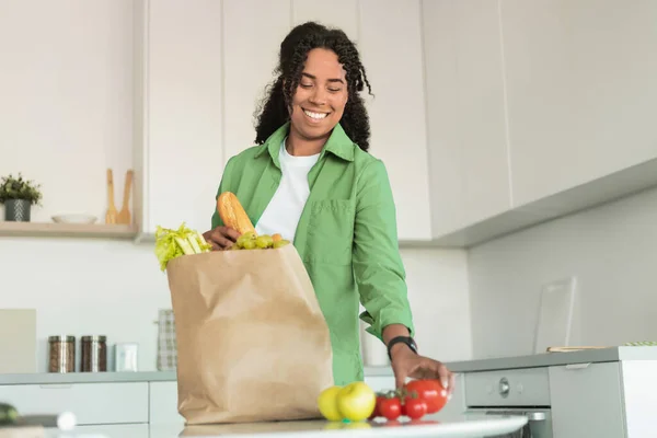 Cheerful black woman in green shirt happily unpacks paper bag of groceries in her kitchen interior, enjoying freshness of food from online shopping spree, ready to cook healthy dinner
