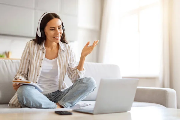 Video Call. Smiling Young Woman Wearing Headphones Teleconferencing On Laptop At Home, Happy Millennial Female Talking And Gesturing At Computer Web Camera While Sitting On Couch, Free Space