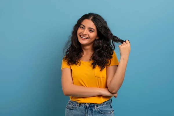 かわいい美しい若いインドの女性は黄色のTシャツとジーンズを着て彼女のカーリーヘアで遊んで カメラで微笑んで 青いスタジオの背景に隔離されたポーズ — ストック写真