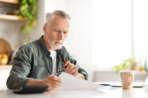 Caucasian senior gentleman reading papers while sitting at desk in kitchen, concentrated elderly male reviewing financial documents, checking insurance contract, accounting bills at home, copy space
