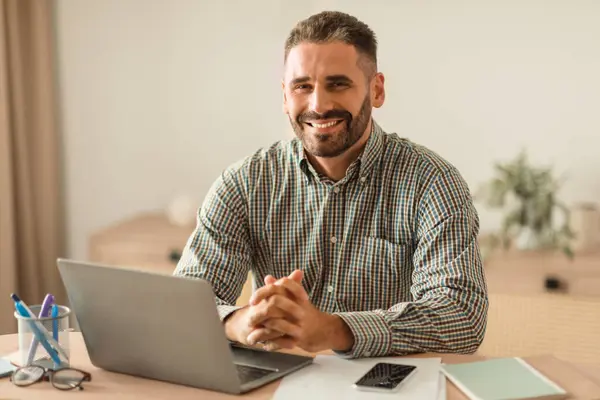 Caucasian middle aged man smiling as working on laptop, posing at work table in modern home office interior, managing his career online. Successful remote work and freelance