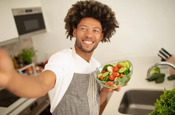 African American guy in apron posing with salad bowl for selfie while cooking in modern kitchen, sharing his culinary hobby online in social media blog, smiling to camera