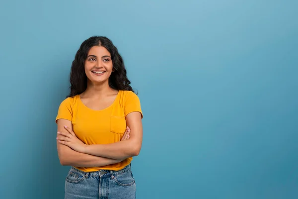陽気なカーリー若いインドの女性 着用ジーンズと黄色のTシャツ コピースペースを見て胸に交差し 素敵なオファーをチェック ブルーの背景に隔離 — ストック写真