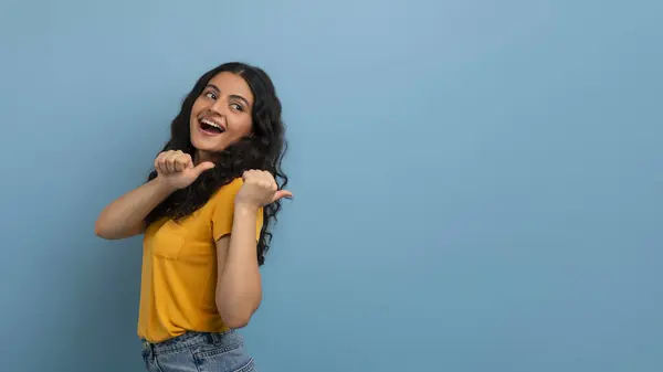 身穿牛仔裤和黄色T恤的积极的卷曲的印度年轻女子20多岁 手指和微笑指向复制的空间 显示出很好的主动表示 孤立在紫色背景 — 图库照片
