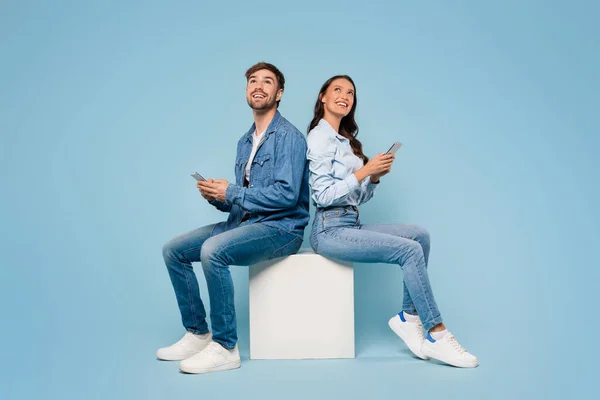 stock image Seated back to back, young European man and woman use their mobile phones, smiling looking at free space on blue studio background, depicting relaxed connectivity