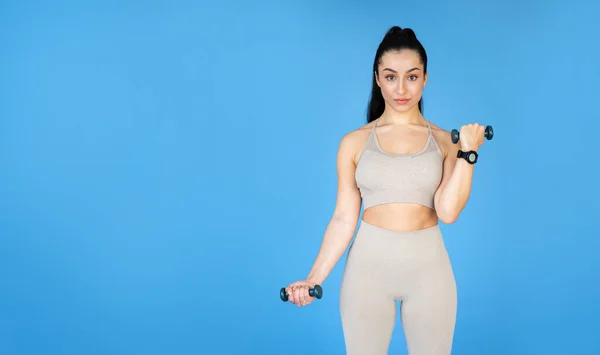 A young well-fit woman is doing strength exercises with weights in front of a blue wall. Shes in exercise clothes, showing her energy and encouragement, empty space for advert next to her