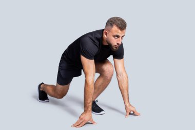 Spor giyim sektöründe güçlü odaklı beyaz erkek sporcu, başlamaya hazır, koşmaya hazır, gri stüdyo arka planında soyutlanmış, tam boy. Vücut bakımı, rekabet, formda ve aktif kardiyo sporu