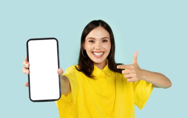 Neşeli, güzel, bin yıllık bir kadın beyaz ekranlı büyük bir telefon gösteriyor ve mavi stüdyo arkaplanı üzerinde başparmak yukarı, akıllı telefon, model, reklam için kopyalama alanı, mobil uygulama, online teklif