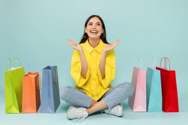 Heyecanlı genç bayan alışveriş meraklısı, renkli alışveriş torbalarının arasında oturuyor, el kol hareketi yapıyor ve gülümsüyor, satış sezonunun keyfini çıkarıyor, kara Cuma anlaşması, mavi arka planda izole edilmiş.