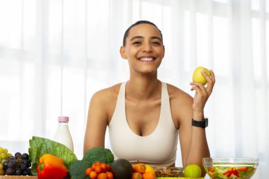 Spor giyim kuşağındaki neşeli Latin bayan açık mutfak içlerinde yeşil elma yiyor, taze sebzelerin ve salatanın tadını çıkarıyor. Diyet, kilo kaybı, sağlıklı yiyecekler, vejetaryen ve vitaminler