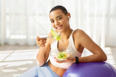 Gülümseyen milenyum Latin kadın spor kıyafetleri içinde, kablosuz kulaklıklar oturma odasının içinde salata yiyor, Fitball 'da öğle yemeğinin tadını çıkarıyor. Evde müzik, sağlıklı yemek ve yaşam tarzıyla beslenme, spor