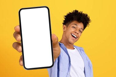 Kıvırcık saçlı neşeli genç bir adam elinde boş ekranlı akıllı bir telefon tutuyor. Uygulama promosyonu için mükemmel, parlak sarı arka plan, model.