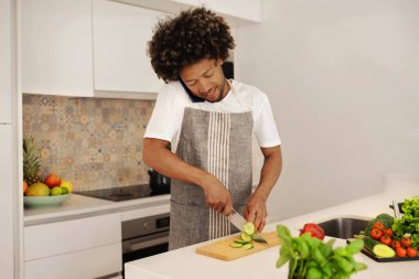Önlüklü genç adam cep telefonuyla konuşur, sağlıklı bir öğle yemeği hazırlar, salata için taze sebze keser, diyet ve refah arasındaki sinerjiyi gözler önüne serer.