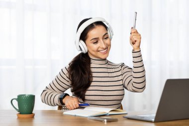 Genç Arap kadın öğretmenin bilgisayarda, kablosuz kulaklıkla çalışmasına sevindim. Oturma odasının içinde el kaldırın. İş, serbest ve çevrimiçi ders, evdeki video görüşmesi.