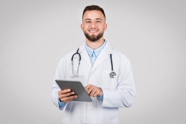 Profesyonel erkek doktor, boynunda steteskop ile, dijital tablet tutuyor ve gülümsüyor, gri zemine karşı modern tıbbi danışmanlığı temsil ediyor.