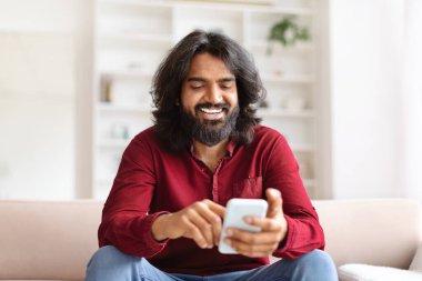 Neşeli, sakallı, genç Hintli bir adam evde koltukta dinlenirken kız arkadaşıyla sohbet ediyor, modern cep telefonu kullanıyor, sosyal medyayı kontrol ediyor, mobil uygulama kullanıyor, yer kopyalıyor.