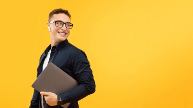 Elinde dizüstü bilgisayarla fotokopi odasına bakan gülümseyen genç adamın portresi, bilgisayar kullanan mutlu adam, sarı stüdyonun arka planında çalışıyor ya da çalışıyor.