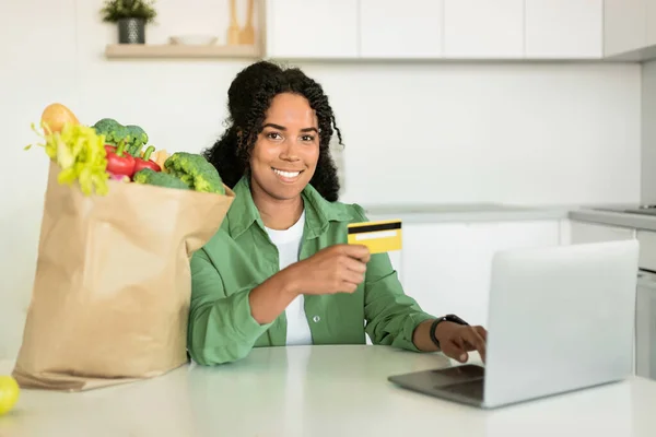 African American lady purchases groceries products online and showing credit card, sitting near laptop computer and paper shopping bag full of fresh grocery items, in her home kitchen
