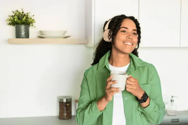 Happy African American Lady Wearing Wireless Headphones Holding Coffee Cup, Enjoying Music And Morning Drink, Spending Time In Domestic Kitchen Indoors. Great Playlist. Copy Space
