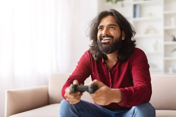 Resting at home, domestic entertainment. Happy young indian man playing video games and smiling while sitting alone on the sofa at home, free space. Modern technologies and entertainment