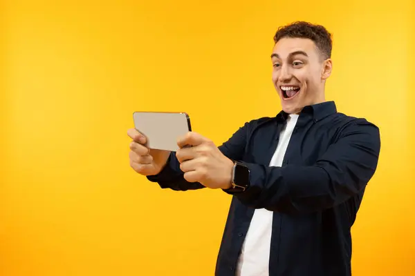 Happy guy have video call with friends or playing online video games, holding smartphone in his hands and smiling, posing isolated on yellow background. Modern gadgets concept