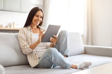 Evinde dijital tablet kullanan güzel beyaz bir kadın, gülümseyen bir kadın, modern bir aletle koltukta dinleniyor, internette geziniyor ya da internetten alışveriş yapıyor, eğlence için teknolojilerden keyif alıyor, kopyalama alanı kullanıyor.