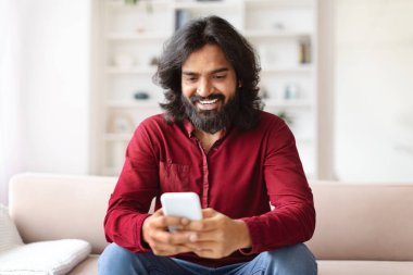 Evdeki koltukta otururken akıllı telefon kullanan mutlu Hintli adam. Cep telefonunda mesajlaşan ya da cep telefonunda yeni uygulama kullanan genç adam, Boşluğu Kopyala