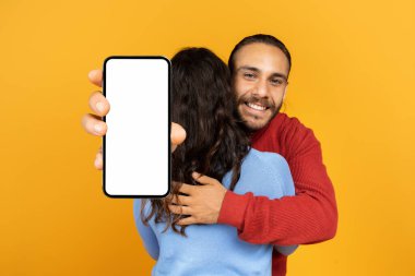 Mutlu yakışıklı adam, sıradan bir şekilde kız arkadaşına sarılıyor, elinde sarı stüdyo arka planında beyaz ekranlı büyük bir akıllı telefon tutuyor, maketini yapıyor. İki kişilik güzel bir anlaşma, harika bir online teklif.