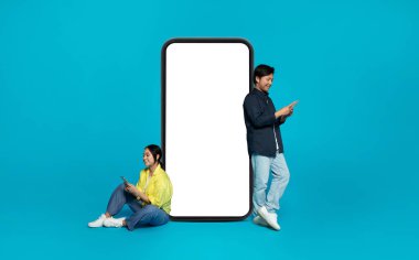 Sıradan giyinmiş mutlu Çinli bir adam ve sosyal ağların yanındaki büyük telefonun yanındaki akıllı telefonlarda sohbet eden mavi stüdyo arka planında izole edilmiş boş ekranlı bir kadın. Çevrimiçi iletişim uygulaması, reklam ve teklif
