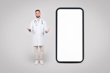 Genç Avrupalı erkek doktor, gri arka plan, model, afiş gibi tıbbi reklamlar için boş ekranı olan büyük cep telefonunun yanında güvenle duruyor.