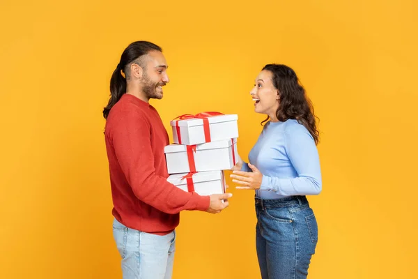 Happy young couple giving gifts to each other, yellow studio background, free space. Loving handsome man giving present to his excited wife or girlfriend. Valentines day celebration, profile side view