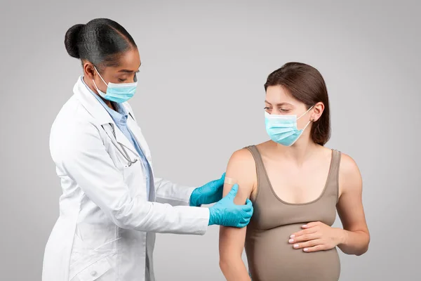 Serious millennial black doctor make injection to pregnant patient during pregnancy, puts plaster on hand, isolated on gray background studio. Vaccination for health care, expecting baby
