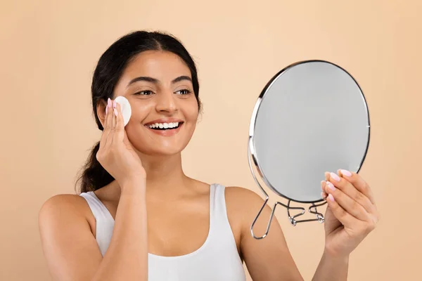 Skin Care Routine. Young Indian Female Holding Magnifying Mirror And Cleansing Face With Cotton Pad, Smiling Eastern Woman Making Beauty Treatments After Shower, Standing On Beige Background