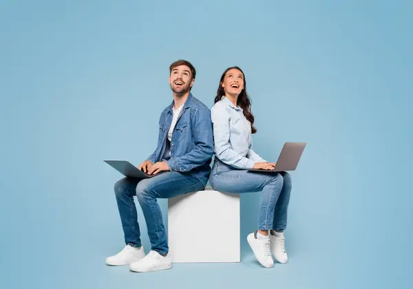 年轻的欧洲夫妇背靠背地坐着 沉浸在膝上型计算机中 眼睛向上看去 在舒适的蓝色画室背景和横幅上看到了自由的空间 — 图库照片