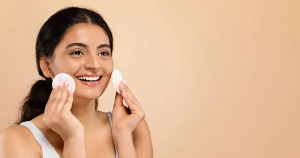 Facial Beauty Treatment. Happy Young Indian Woman Using Cotton Pad For Cleansing Face, Beautiful Eastern Female Looking Aside At Copy Space While Standing Over Beige Studio Background, Panorama