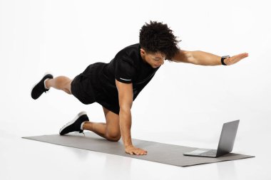 Atletik genç adam dizüstü bilgisayarın önünde çevrimiçi pilates kursuna katılıyor, not defterindeki rutin antrenman ipuçlarını kontrol ediyor, beyaz stüdyoda paspas üzerinde egzersiz yapıyor, dört ayaklı çapraz bacak uzatma hareketi yapıyor.
