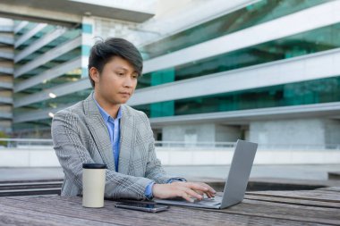 Çevrimiçi çalışan Asyalı girişimci, ofis binasının yanında oturan, kentsel alandaki e-öğrenim ve serbest iş görevlerine odaklanmış, bilgisayar hakkındaki dijital raporları inceliyor