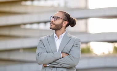 Takım elbiseli, kollarını çapraz yapmış, açık havada poz veren, modern iş merkezinin yanında duran gözlüklü, yakışıklı, yeni yetme erkek girişimci, fotokopi uzayına bakıyor.