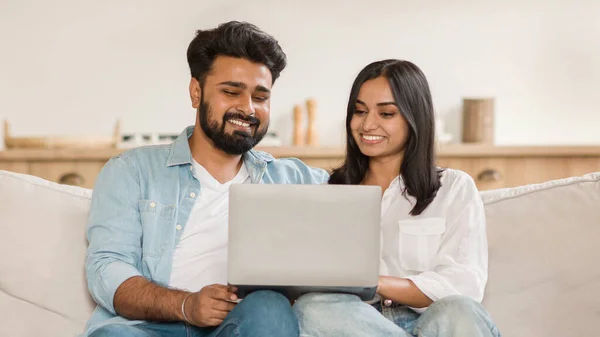 幸せなインドのカップルは 快適にソファに座って ノートパソコンでコンテンツを見ることに従事し カジュアルな設定で喜びとトータルさを放射します リビングルームのインテリア — ストック写真