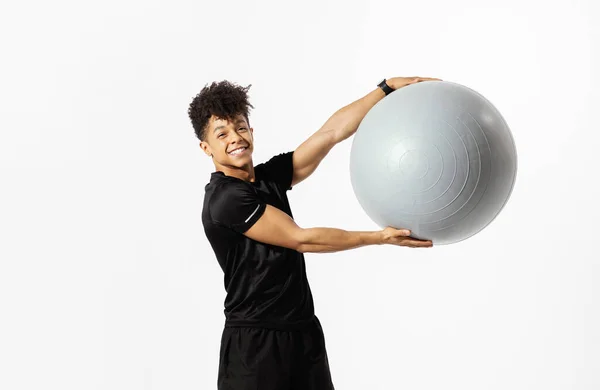微笑年轻的拉丁小伙子举起适形球微笑着对着相机 同时在白色工作室的背景下锻炼 穿运动服的有决心的男人练体操 做有氧运动 — 图库照片